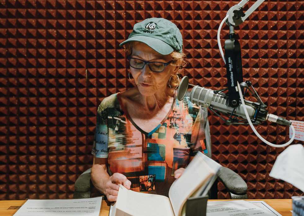 A woman seated in a recording booth intently focuses on the open book in front of her, as she reads into an ElectroVoice microphone.