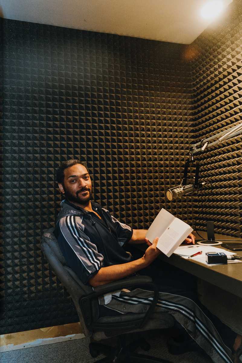 A man looks towards us as he sits in a recording booth with a novel open in his hands.