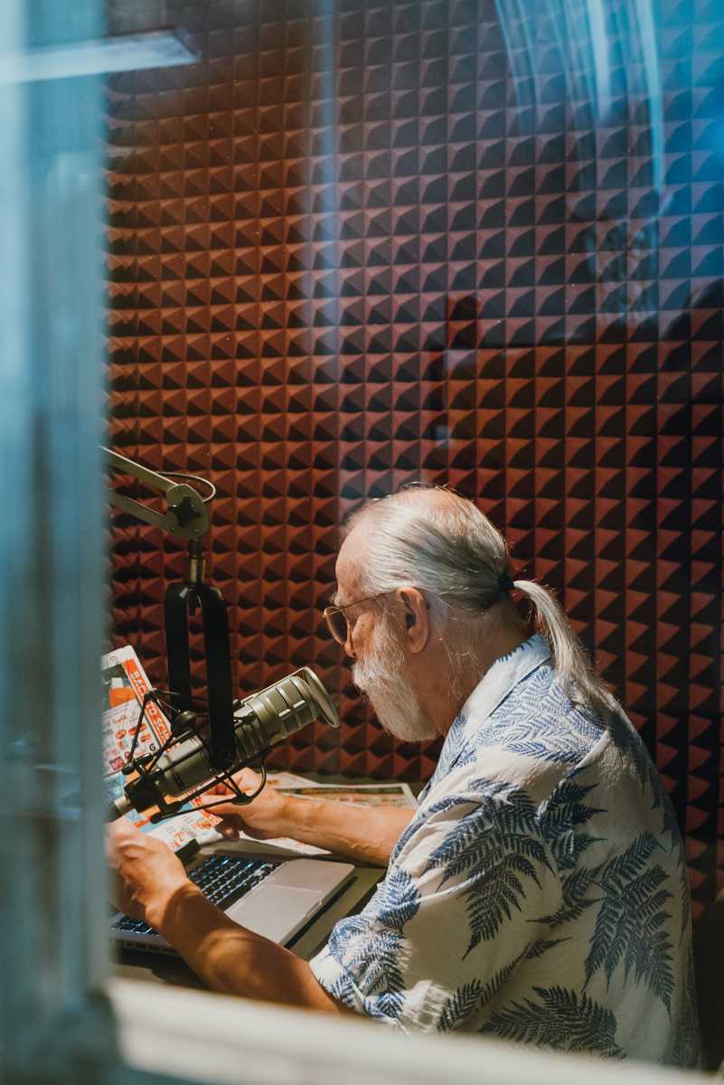 A man sits in a recording booth reading from the colorful newspaper he holds in his hands.