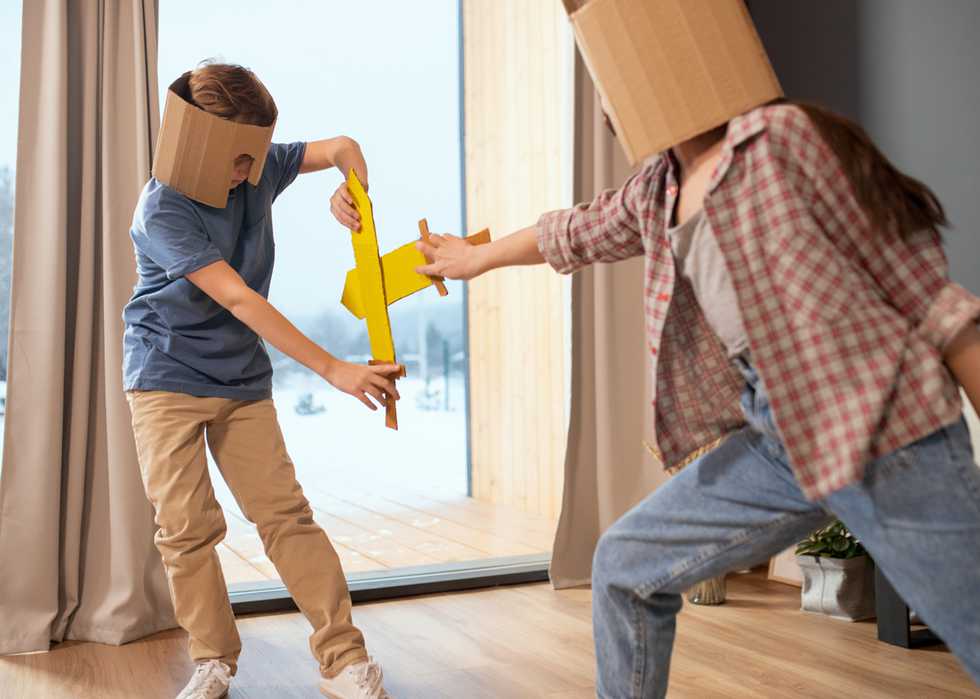 Two contemporary kids in cardboard helmets fighting with toy swords at home